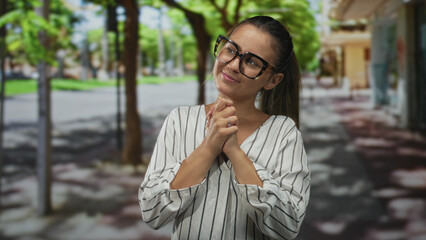Fototapeta premium Young woman with clasped hands and gentle smile on a city street lined with trees and storefronts; hopeful gratitude.