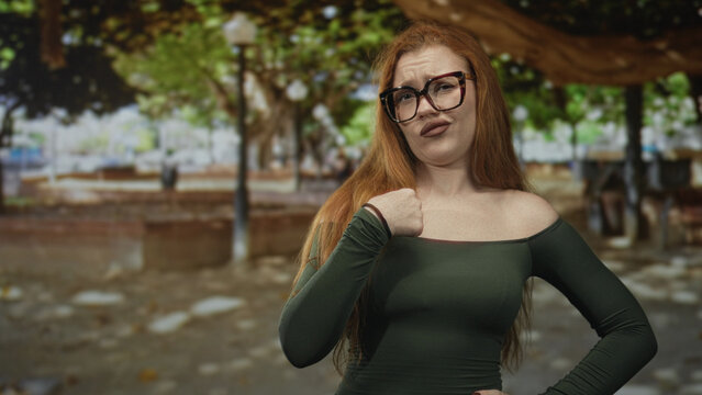 Redhead woman wearing glasses points index finger with pursed lips in a street park scene, off shoulder top visible; warning disapproval.