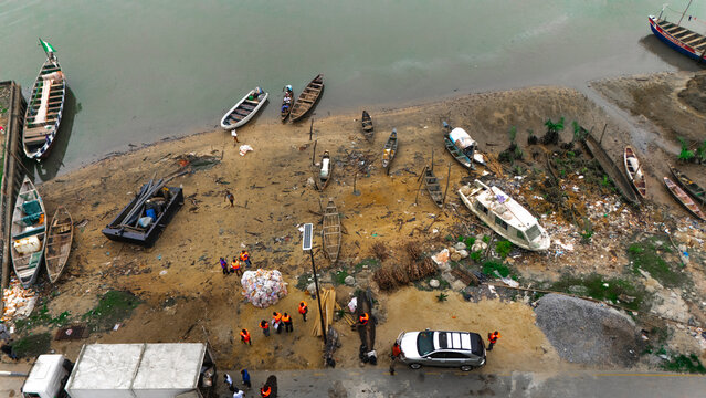 Aerial view of a riverbank with various wooden boats, small vessels, and people working near piles of debris and parked vehicles in Rivers, Nigeria.