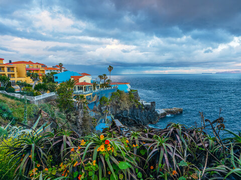 Colorful residential houses on a coastal hill looking over a dramatic dark stormy ocean under a cloudy sky in Canico, Madeira, Portugal.