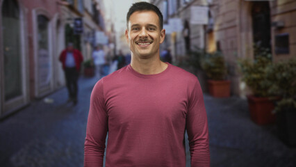Obraz premium Man with mustache turning head and smiling while wearing a crewneck shirt on a narrow cobblestone street lined with shops; friendly confidence.