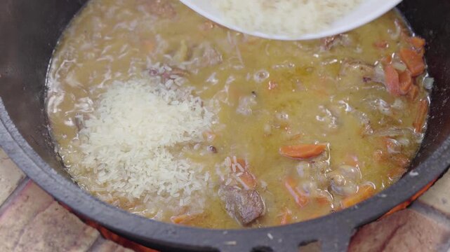 chef adding white rice from a plate into a boiling traditional cast iron cauldron filled with hot meat, carrots, and onions broth to cook Uzbek plov over an open fire.