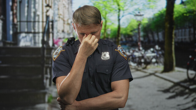 Police officer man pinches nose by visible badge on street with folded arms and navy uniform shirt; stress reflection.