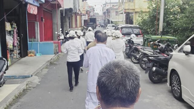 A rear view of a community procession featuring individuals dressed in matching white uniforms and hats walking down a narrow urban street lined with vehicles and shops.