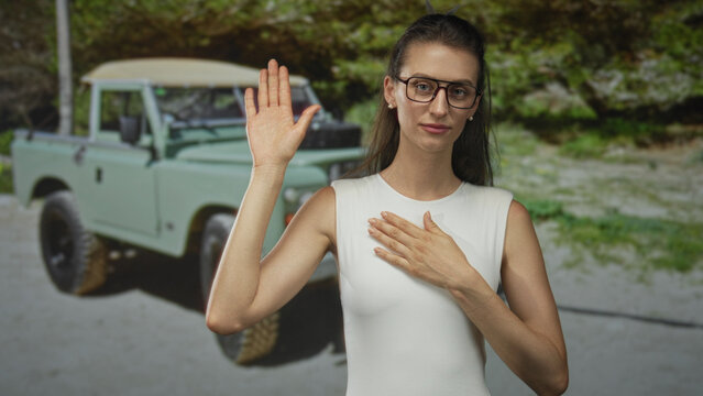 Woman raises hand with other hand on chest beside a green offroad vehicle in forest wearing white sleeveless garment; oath sincerity.