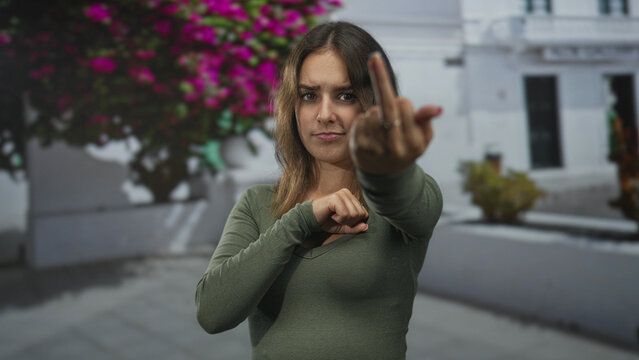 Young hispanic woman shows middle finger to camera in a city street framed by white building and pink bougainvillea; defiance.
