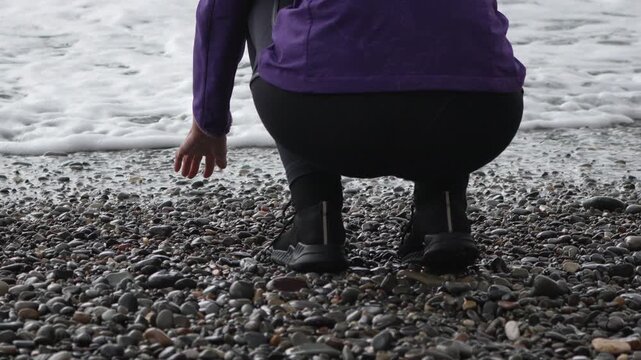 Squatting, beach, pebbles, woman crouches down on shingle shore near breaking ocean waves.