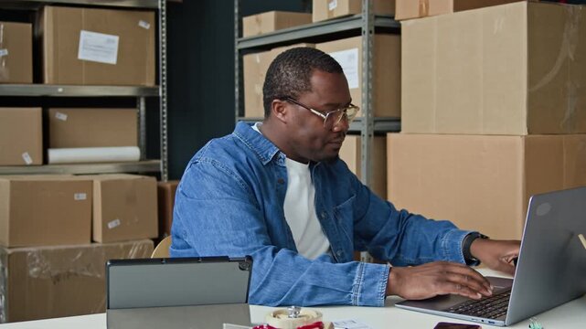 Arc shot of African American young man scanning barcode on cardboard box with scanner and typing on laptop in warehouse. Clip is perfect for e-commerce logistics and inventory management promotions