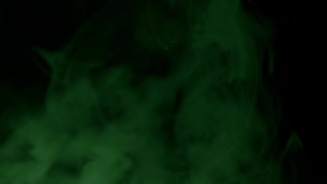 Thick clouds of yellow green smoke drift slowly and morph into teal blue hues against a black background. Concept of toxic fumes, chemical reactions, Halloween mystery, and magical spells.