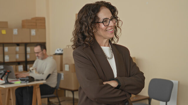 Woman smiling with arms crossed and parcels and laptop amid stacked boxes on shelves in building; confidence entrepreneurship.