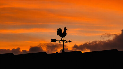  A striking silhouette of a traditional rooster weathervane stands atop a rooftop against a dramatic sunset sky filled with rich orange, amber, and gold tones. © cratervalley