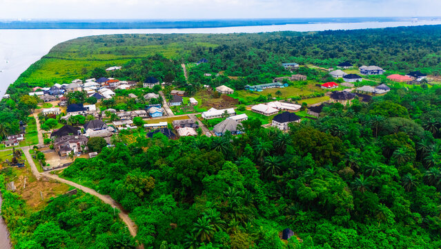 Aerial view of a small settlement nestled within dense tropical forest along a wide river, featuring scattered houses with varied roof colors in Minima, Rivers, Nigeria.
