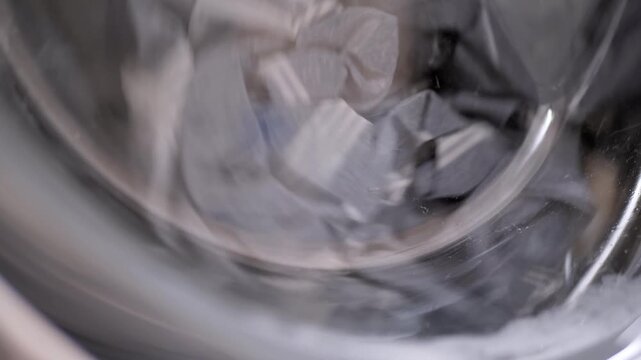Wet garments tumble inside a spinning washing machine drum with splashing soapy water. Concept of household cleaning, daily domestic chores, and modern home appliance usage.