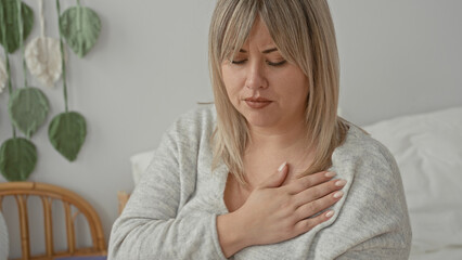 Fototapeta premium Woman, blonde, clutching chest with hand on chest gesture suggesting heartattack while seated on bed beside a book and rattan chair in studio; concern health awareness.
