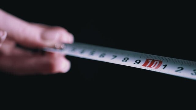 A human hand pulls a metal tape measure forward against a completely black background with focus on the numbers. Concept of precision, accurate measurement, and construction planning.