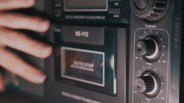 A close-up of a female hand touching an old, retro radio cassette player. The shot focuses on the tuning and volume knobs of the vintage boombox. Concept of nostalgia, 80s music, and