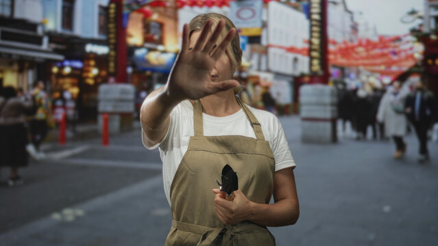 Young woman gardener holding pruning shears with palm out in stop gesture blocking the camera on a crowded street; defiance boundary.