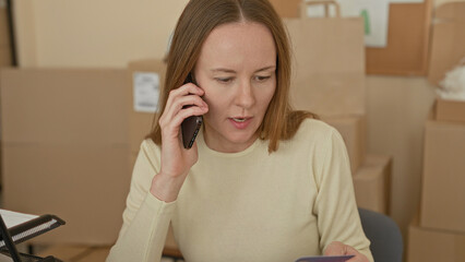 Fototapeta premium Woman holds smartphone to ear and checks creditcard while seated among stacked cardboard boxes and shipping labels in building; concern logistics.