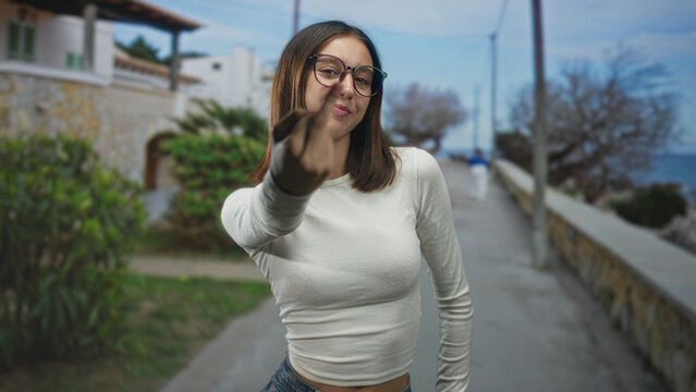 Young woman in white shirt flips middle finger to camera on a seaside street promenade, hand extended and smirking; rebellion defiance.