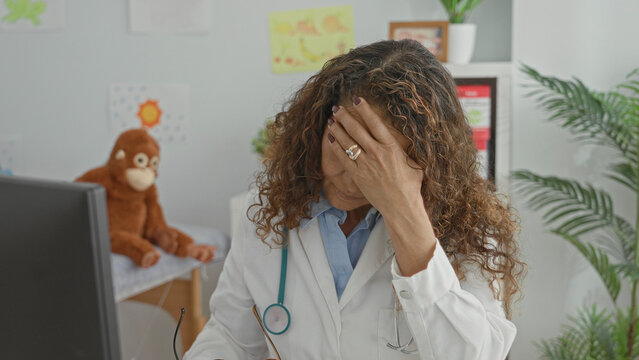 Woman doctor with stethoscope covering forehead with hand and holding glasses at computer desk in pediatric clinic; fatigue.