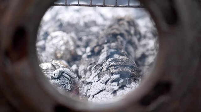 Glowing wood embers and charcoal burn intensely within a round metal firebox, radiating warmth and heat. This scene evokes concepts of cozy outdoor cooking, camping, and the fundamental