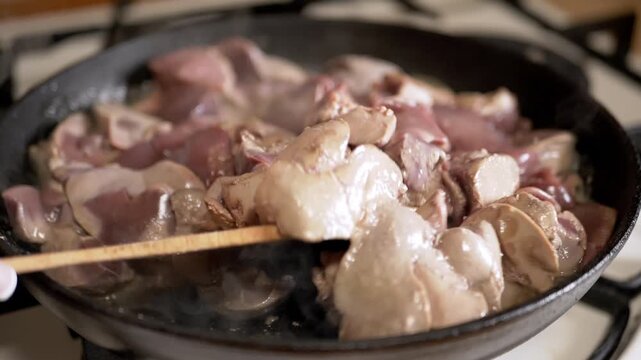 Raw chicken livers are tossed with a wooden spatula in a hot cast iron skillet on a kitchen stove. Concept of traditional cooking, poultry preparation, and nutritious offal recipes.