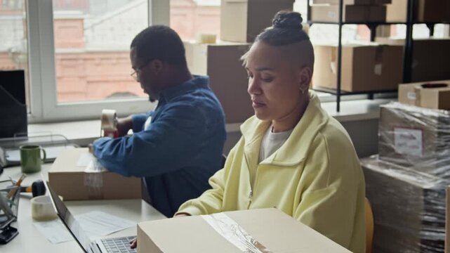 Handheld shot of African American man and diverse female colleague packing cardboard boxes and using laptop in warehouse. Footage is ideal for e-commerce, logistics, and business shipping concepts