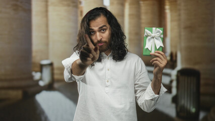 Fototapeta premium Man young arab hispanic with beard and long hair holds green gift box with white bow and points finger to it on old town street; skepticism.