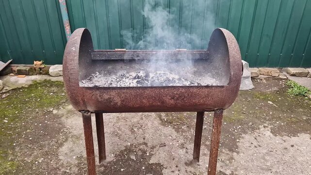Smoke rises from hot gray ash inside a rusty DIY metal barbecue grill made from a repurposed tank. Concept of outdoor cooking, rural lifestyle, and rustic backyard barbecue preparation.