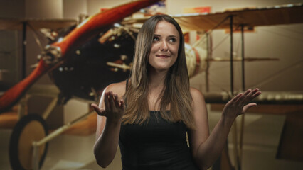 Brunette young woman in black tank top with palms up gesture in front of vintage biplane inside studio set  playful curiosity. © Krakenimages.com
