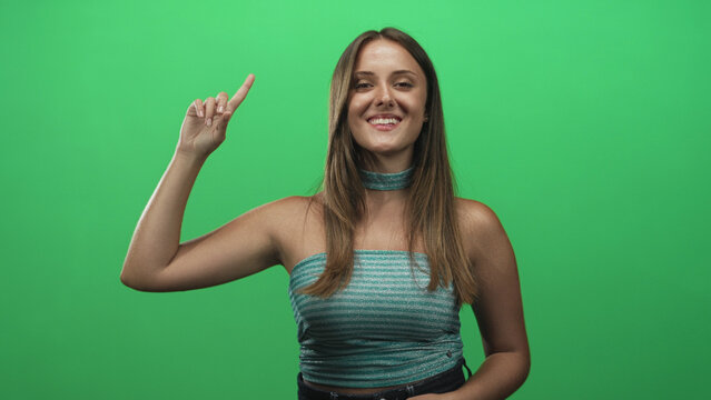 Woman points index finger up in studio green screen while smiling, finger raised and arm slightly bent; confidence.