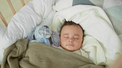 Obraz premium Baby boy sleeping with closed eyes in a crib, face visible, swaddled under a green blanket beside a plush rabbit inside a building; serene rest.