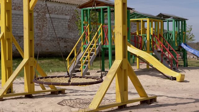 An empty bright yellow nest swing hangs on a colorful wooden playground set built on a sandy surface. Concept of outdoor childhood recreation, summer playtime, and neighborhood parks.