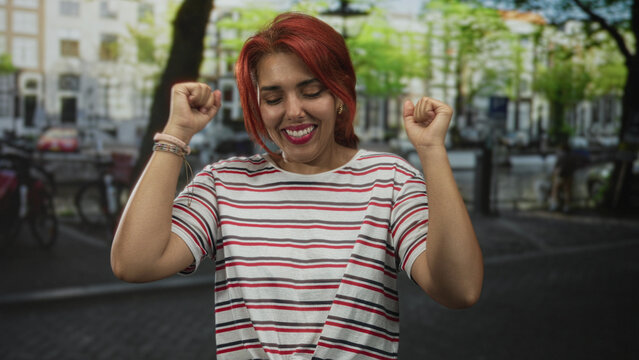 Woman with hands clasped in prayer on a cobblestone street, striped shirt and red hair, eyes closed and smiling, wrists showing watch and bracelet; gratitude.