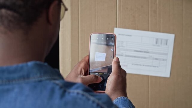 Over the shoulder view of African American young man scanning shipping label on cardboard box with smartphone in warehouse. Clip is ideal for e-commerce logistics, inventory tracking, and delivery ads