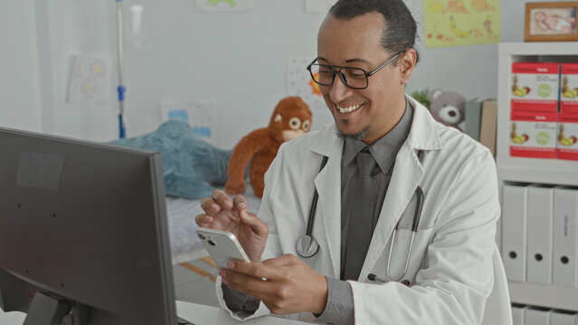 Man doctor in clinic taps smartphone with index finger while stethoscope rests on his neck and computer monitor shows records; friendly care telemedicine.