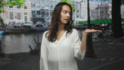 Fototapeta premium Woman in white blouse holds palm up, presenting on a street by a canal with bicycle and boats visible; curiosity.