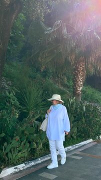 Senior woman walking outdoors near palm trees, cactus plants and lush greenery on a sunny day. Active aging and independent senior lifestyle concept.