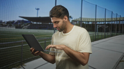 Fototapeta premium Hispanic man with beard typing on laptop, holding device with both hands on a street sidewalk beside a stadium fence and bleachers; productivity.