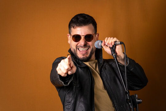 Hombre rockero con chaqueta de cuero y lentes cantando en&eacute;rgicamente con su micr&oacute;fono y se&ntilde;alando a c&aacute;mara