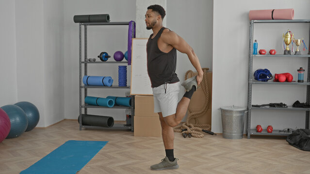 Man in studio holds ankle for standing quad stretch beside exercise mat and shelving with ropes and dumbbells; determination fitness.