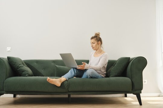 Middle-aged woman is seated on a green couch, engaged with her laptop in a well-lit living room. She appears focused and relaxed, with natural light illuminating the space