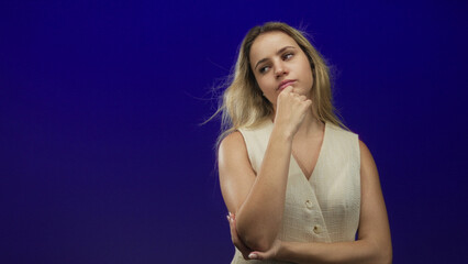 Naklejka premium Woman young blonde touching chin with hand, thoughtful pose in studio against deep blue backdrop; contemplation curiosity introspection.