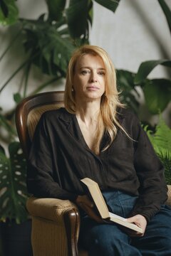 A woman with long blonde hair sits comfortably in a vintage armchair holding a book. She is surrounded by vibrant greenery, creating a serene and inviting atmosphere perfect for reading