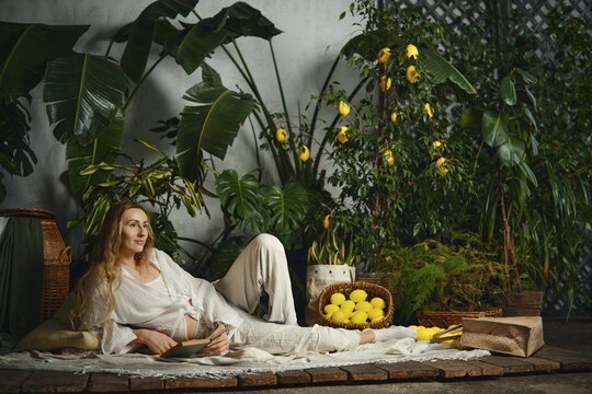 Dreamy woman sits on the floor surrounded by green plants and yellow lemons. She holds a book while resting on soft fabrics. Natural light comes in, creating a warm atmosphere and inviting scene