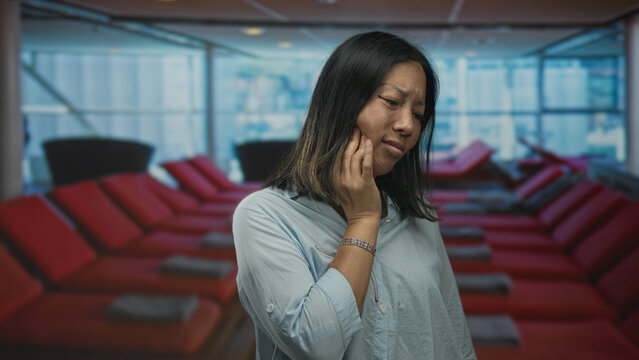 Woman with hand on cheek in lounge building by red loungers, touching jaw in mild pain; toothache concern.