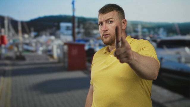 Man in yellow polo points finger to camera with stern face at port dock beside moored boats and pilings; warning confrontation admonition.