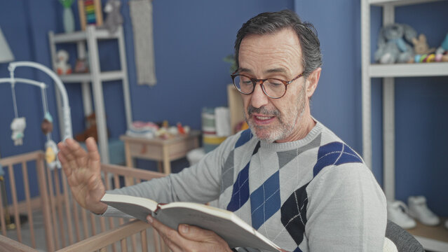 Man reading book with hand resting on cradle in a baby room building, wearing glasses and sweater; serenity.