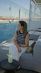 Obraz premium Woman sipping cocktail on cruise terrace, hand holding glass with pineapple garnish and sunglasses on head while seated on sofa; summer getaway relaxation.