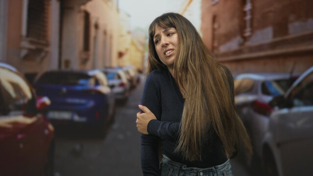Woman clutching her shoulder with bare hand on a narrow urban street, long hair, eyes closed and winced face amid parked cars; discomfort.